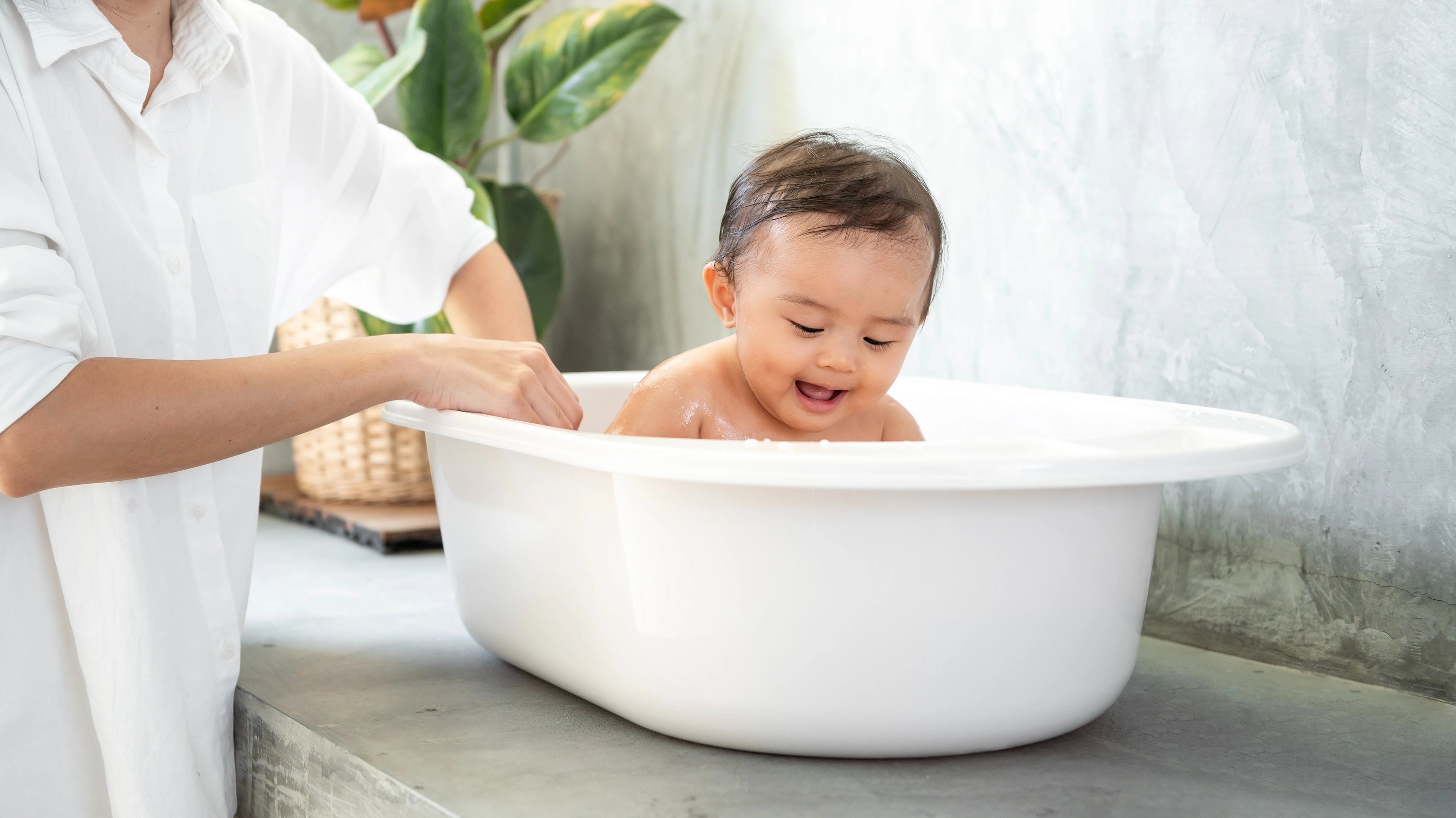Baby being bathed in a white basin by an adult in a home setting.